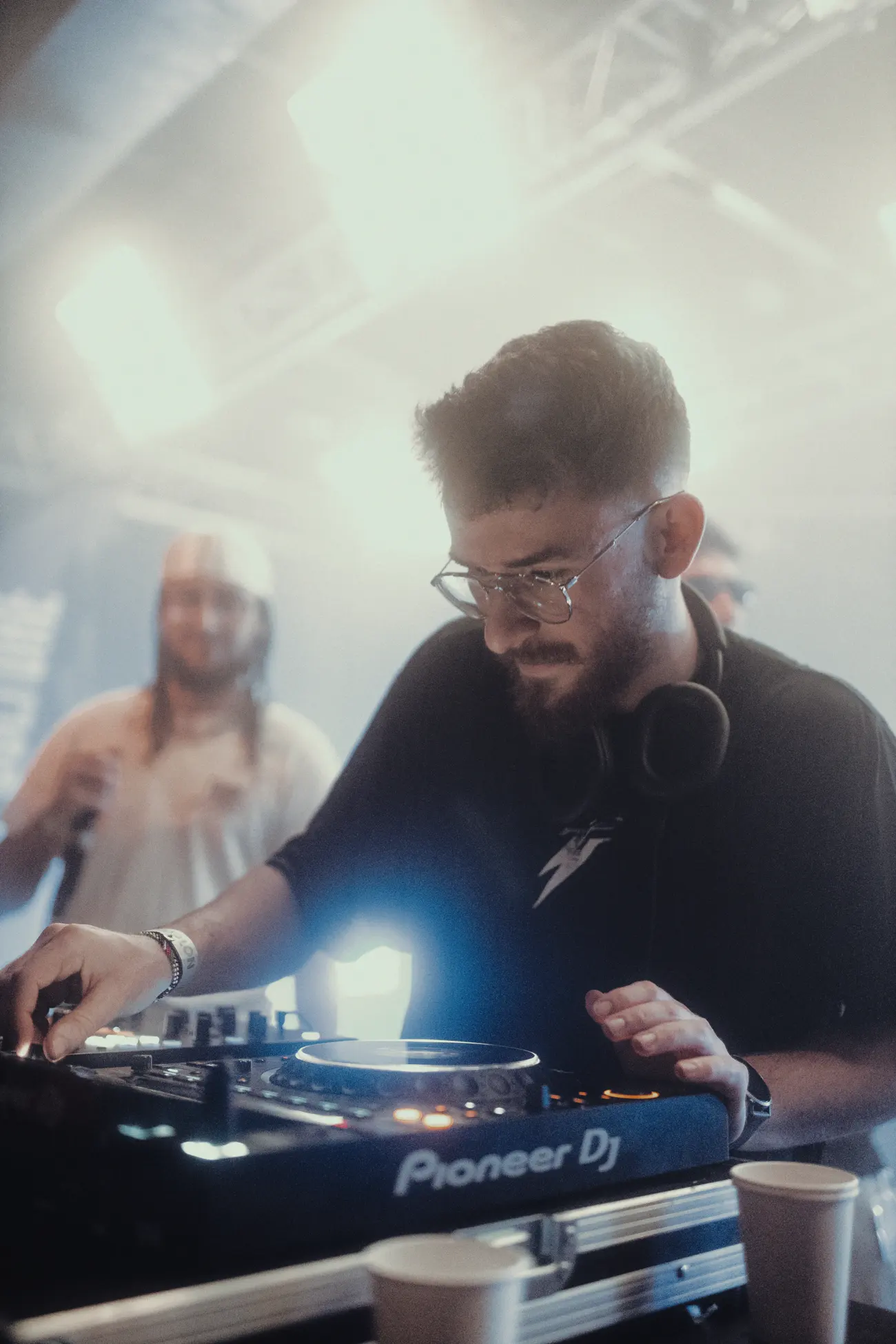 A DJ mixing under white light, the room hazy behind.